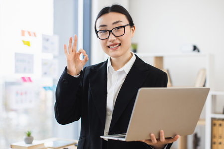 Confident Asian businesswoman in formal wear holding laptop, making okay gesture. Young female professional expressing approval and success in modern office environment.の写真素材