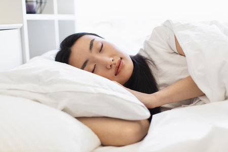 Asian woman asleep in cozy bed, surrounded by white linens, capturing serenity and tranquility of restful sleep. Perfect depiction of relaxation and peaceful atmosphere.の写真素材
