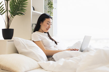 Asian woman in her 20s using laptop in cozy bedroom setting. Relaxed and focused atmosphere with natural daylight and indoor plants adding calmness to home workspace.の写真素材