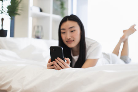 Asian woman in twenties lying on bed using smartphone. Relaxed atmosphere, connected with digital world, enjoying leisure time. Bright room, peaceful setting, modern lifestyle conveyed.の写真素材