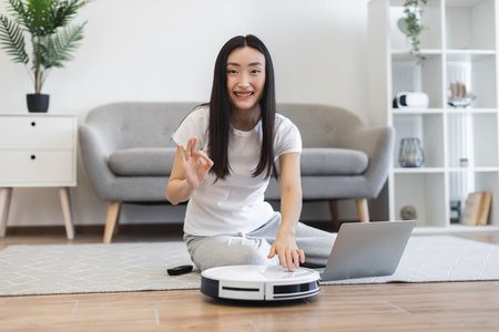 Young Asian woman in casual attire uses robot vacuum cleaner in modern living room. She operates device while seated on floor near laptop, enjoying convenience of smart technology.の写真素材