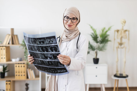 Young Middle Eastern female doctor wearing hijab examining x-ray in medical office setting,...