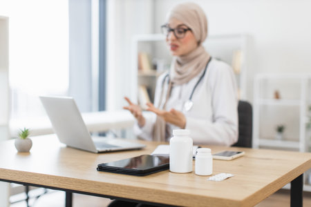 Middle-aged Muslim female doctor having video call with patient, using laptop in bright office. Medical supplies on desk suggest virtual consultation or telehealth services.の写真素材