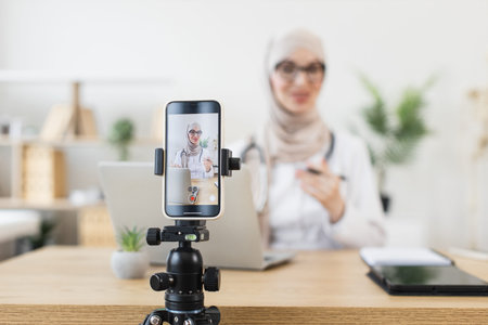 Young Muslim woman recording lesson on smartphone in bright home office. Middle Eastern female...