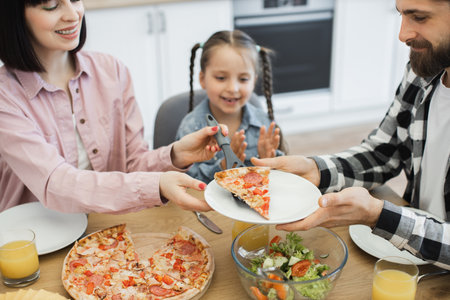 Caucasian family with parents and young daughter enjoying pizza breakfast at home kitchen table, sharing happy moments and smiles.の写真素材
