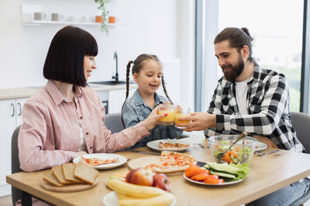 Happy Caucasian family with young parents and daughter clinking glasses of juice during breakfast in bright kitchen. Fresh fruits and healthy food create joyful atmosphere.の写真素材