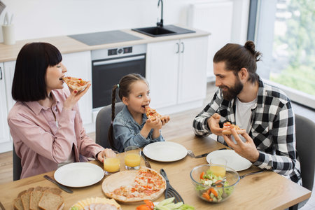 Caucasian family of three, parents and child, sharing pizza for breakfast at home kitchen table. Joyful morning scene with family interaction and delicious food.の写真素材