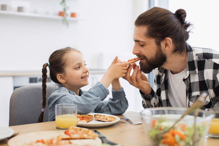 Caucasian father and daughter sharing pizza breakfast. Smiling girl with braided hair feeds bearded dad. Bright kitchen atmosphere with natural light, orange juice, and salad on table.の写真素材