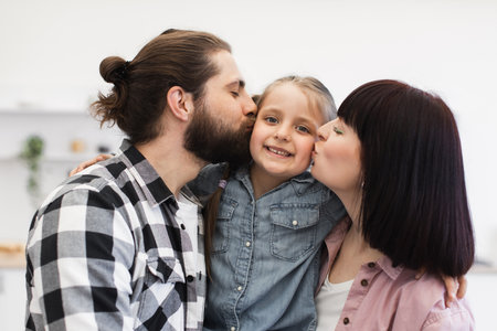 Caucasian mom and dad lovingly kiss young daughter in bright kitchen setting, showcasing joy and togetherness.の写真素材