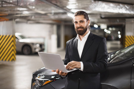 Caucasian male businessman in suit using laptop in modern underground parking garage setting....