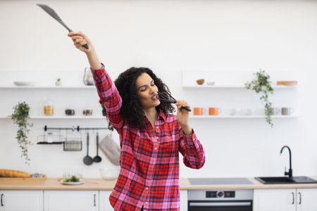 Smiling African woman in twenties holding kitchen tools, singing in modern bright kitchen setup....