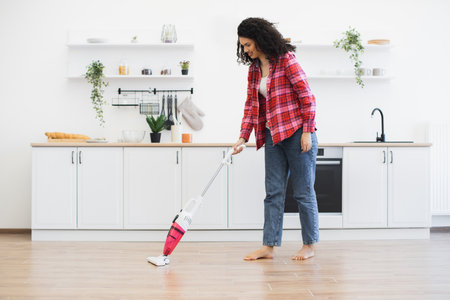Young African woman using cordless pink vacuum cleaner barefoot in bright kitchen. Domestic activity.の写真素材