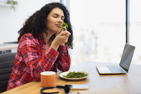 Young African American woman sitting in office eating salad while enjoying fragrance. Relaxed moment during work with plate of healthy food, coffee cup, laptop on deskの写真素材