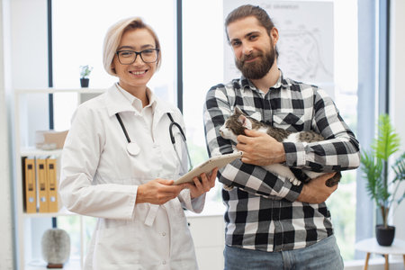 Veterinarian showing medical prescription on tablet to young male owner holding his cat. Indoor veterinary clinic setting featuring tools and trusting interaction promoting careの写真素材