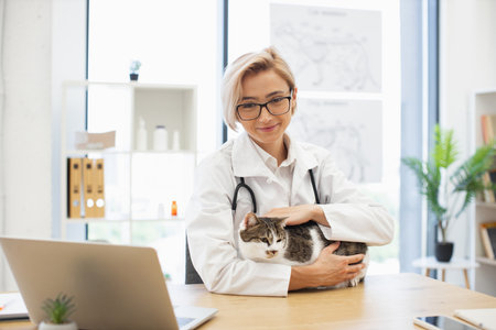 Young adult female veterinarian in white coat with stethoscope caring for domestic cat in clinic. Professional pet healthcare and animal care concept. Bright office setting with plants and equipment.の写真素材