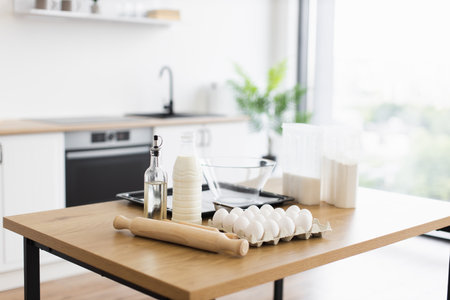Kitchen scene with eggs, milk, flour, and utensils on wooden table in modern interior. Bright natural light through window. Concept of home baking, recipe preparation, and culinary creativity.の写真素材
