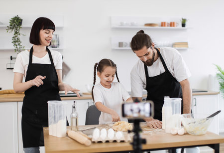 Caucasian family cooking together in kitchen, recording video while bonding over food preparation. Parents and child in aprons kneading dough, smiling and enjoying shared cooking experience.の写真素材