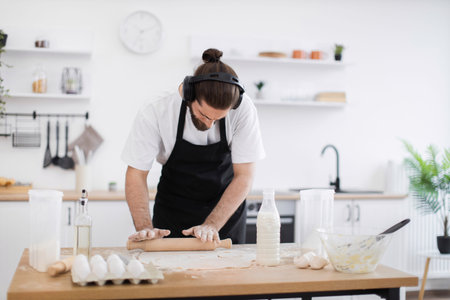 Man wearing headphones and apron rolls dough with rolling pin on kitchen table. Modern kitchen...