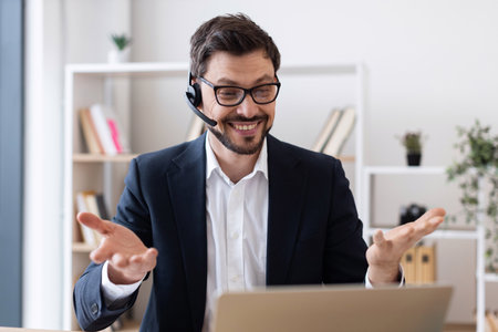 Smiling young man in formal suit using headset and laptop during video call in office. Professional working environment with bookshelves in background showcases cheerful approach to remote working.の写真素材