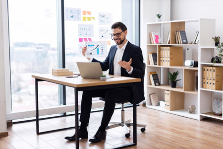 Young adult Caucasian male in suit seated at desk with laptop using graphs and charts for business analysis in bright modern workplace. Office background includes shelves with books and files.の写真素材