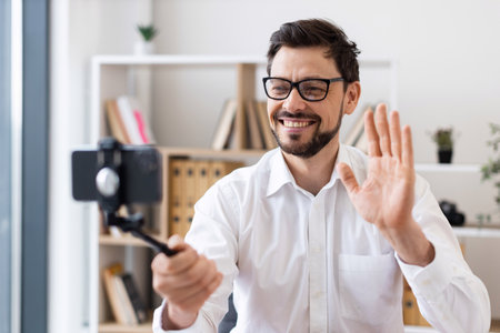 Middle-aged man recording video call with smartphone indoors expressing happiness and engagement. Dressed in white shirt, captured in bright modern office environment.の写真素材