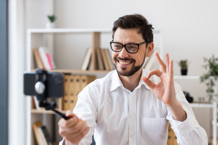 Middle-aged man recording video call with smartphone indoors expressing happiness and engagement. Dressed in white shirt, captured in bright modern office environment.の写真素材