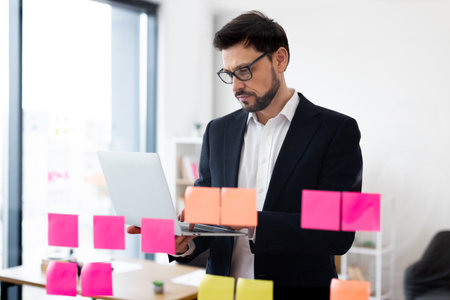 Professional male adult in office setting working on laptop with organized sticky notes for project planning and collaboration. Office layout features modern urban designの写真素材