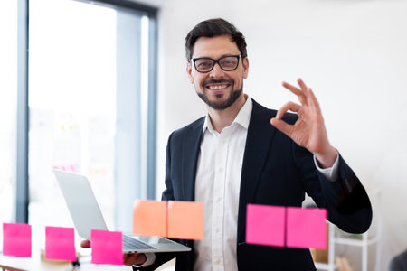 Businessman in thirties wearing glasses and suit using laptop managing tasks on transparent glass board, smiling confidently, office environment.の写真素材