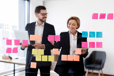Business colleagues, young man and woman, collaborating on project ideas, using colorful sticky notes on transparent board while smiling in office environmentの写真素材