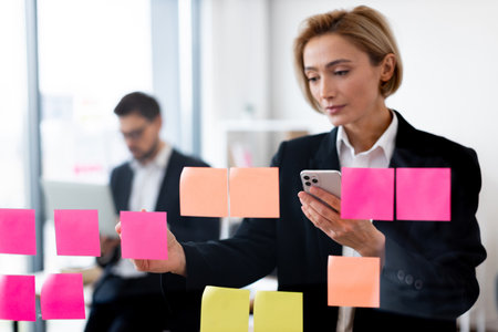 Professional adult caucasian businesswoman interacting with colorful sticky notes on glass board while man working in background in modern office environmentの写真素材