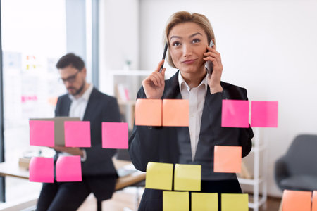 Professional businesswoman multitasking with phones and managing tasks using colorful sticky notes on transparent board. In the office setting, male colleague collaborating in background.の写真素材