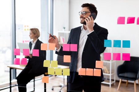 Male adult wearing suit using sticky notes on glass for organization while on phone call. Professional working environment with woman colleague in background using laptopの写真素材