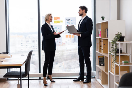 Two business professionals, one male and one female, discussing work in an office. Both are wearing formal attire, standing near a large window, with charts and diagrams visible in the background.の写真素材