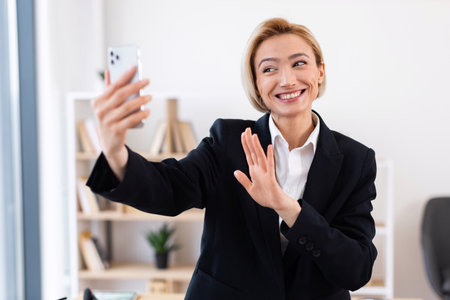 Smiling businesswoman in formal black suit conducting remote video call in bright office environment. Representing concepts of communication, technology, and professional interactions.の写真素材