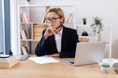 Businesswoman with glasses and blonde hair sitting at desk with laptop. Middle-aged professional in office setting focused on work. Concept of leadership productivity career and workplace.の写真素材