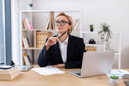 Middle-aged businesswoman in formal suit sits at desk contemplating work ideas with laptop.の写真素材