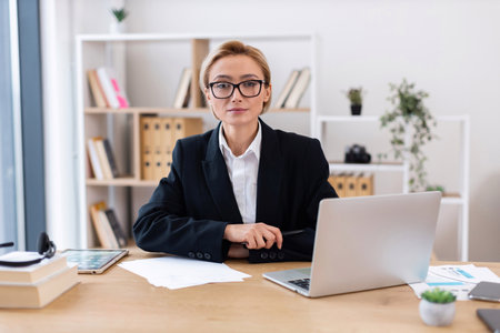 Confident adult woman in formal attire working in modern office environment. Professional focus with laptop, papers, books emphasizing business, professionalism, and productivity. Bright workspace.の写真素材