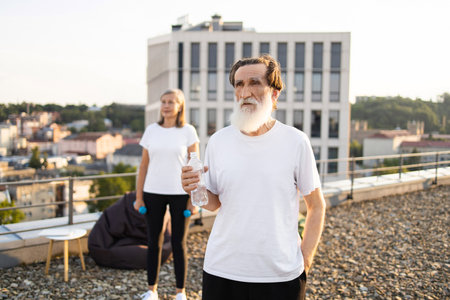 Elderly man with white beard holding water bottle, woman wearing white shirt, outside on rooftop. Scenic urban background, focus on healthy fitness routine, water hydration during break.の写真素材
