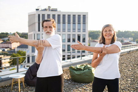 Active senior man and woman enjoying arms stretch exercise during yoga session outdoors. Both wearing casual activewear. Background features city skyline and urban setting, conveying health awarenessの写真素材
