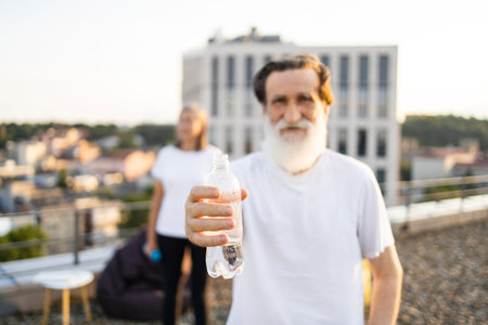 Elderly man with beard dressed in white shirt on urban rooftop holding water bottle. Background features a blurry cityscape. Scene exudes fitness, hydration, maturity, and enjoying active lifestyle.の写真素材