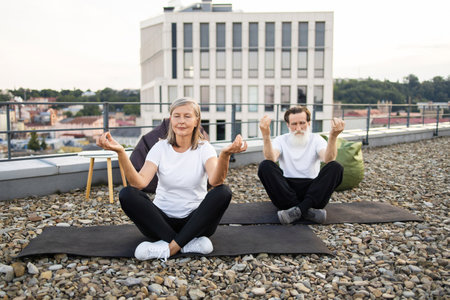 Elderly man and woman sitting in lotus pose practicing meditation on rooftop urban area.の写真素材