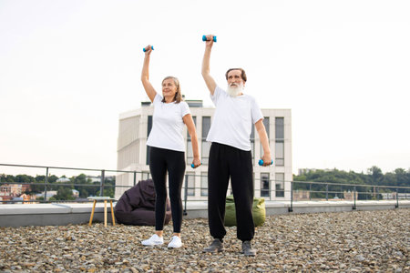 Senior man and woman performing fitness exercise with dumbbells on rooftop terrace outdoors under open skyの写真素材