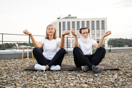 Senior man and woman practicing meditation together, sitting in lotus pose on rooftop stones....