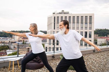 Senior man and woman practicing yoga in outdoor rooftop session overlooking urban landscape. Both wearing casual tops and comfortable clothing while performing warrior yoga poses, emphasizing balance.の写真素材