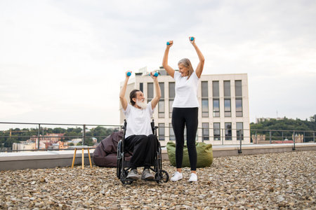 Senior man in wheelchair and woman exercising with dumbbells outdoors on urban rooftop promoting active lifestyles. Encouragement, togetherness, health, vitality, and support in fitnessの写真素材