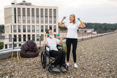 Senior man in wheelchair exercising with dumbbells assisted by woman on building rooftop. They are enjoying fitness outdoors together, focusing on health and rehabilitation in inclusive environment.の写真素材