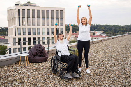 Senior woman and man, one in wheelchair, exercising together outdoors using dumbbells, showcasing fitness and support.の写真素材