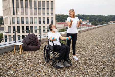 Senior man in wheelchair exercising with dumbbells alongside supportive woman on rooftop. Scenic urban backdrop emphasizes health and strength for elderly couples.の写真素材