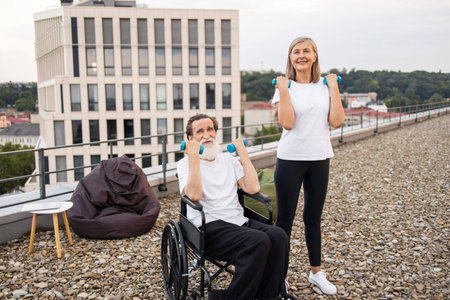 Senior man in wheelchair exercising with dumbbells assisted by woman, both smiling outdoors.の写真素材