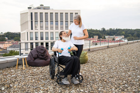 Elderly couple exercising together outdoors. Senior woman supporting her husband seated in...